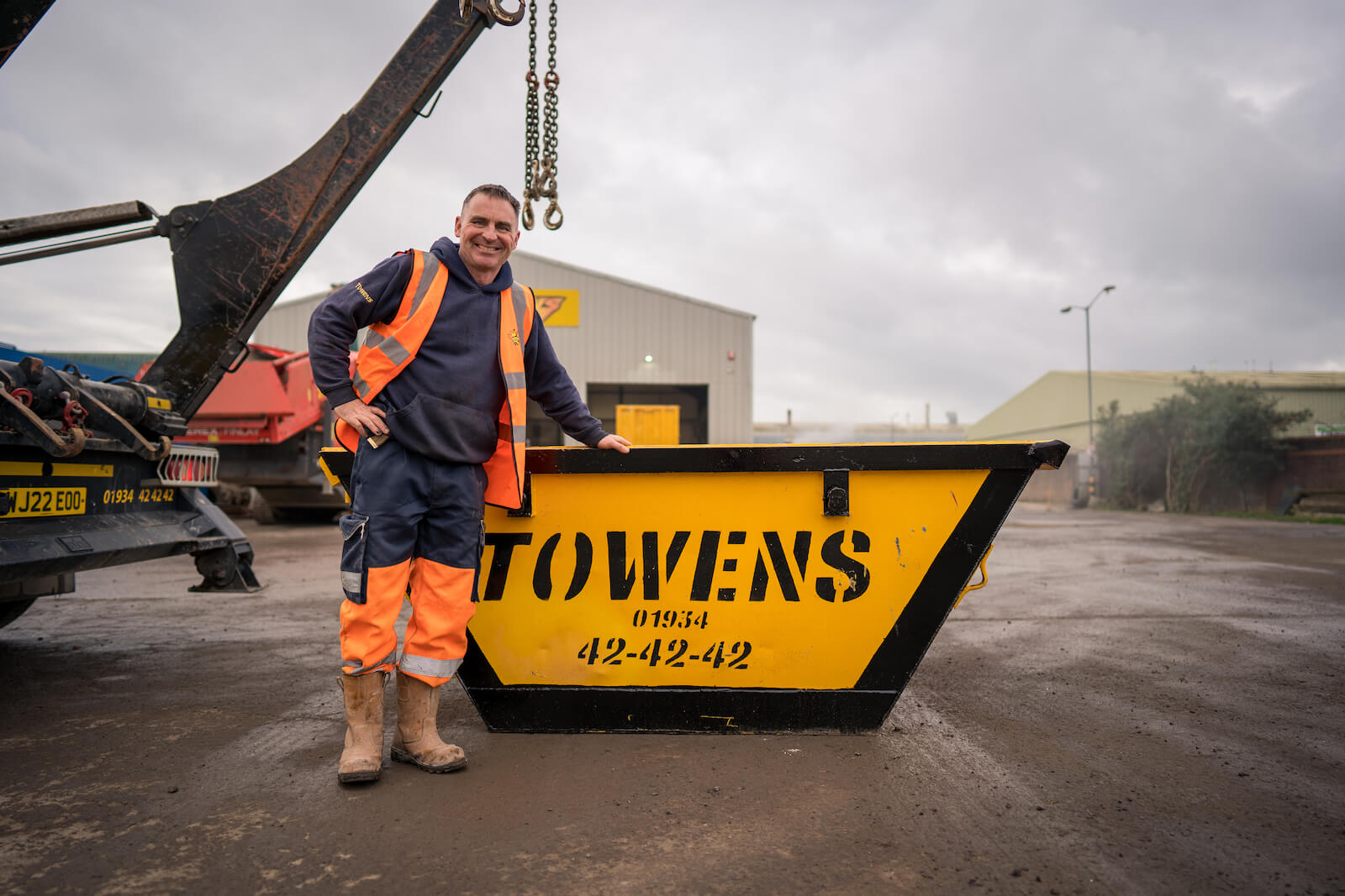 Towens employee in high-visibility gear standing beside a yellow 2-yard skip branded with Towens contact details, in an industrial setting.