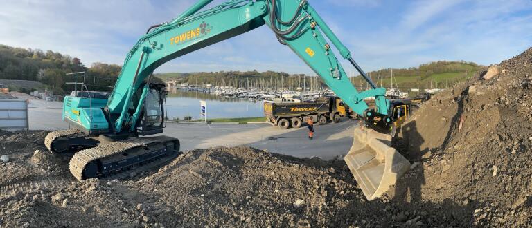 Towens excavator loading aggregate into a tipper lorry at a construction site near a marina, with additional plant and vehicles in view.