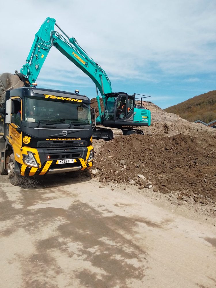 Towens excavator loading soil and rubble into a Volvo FM tipper lorry at an active construction site.