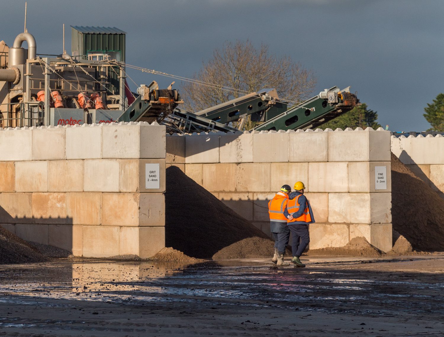 Two construction workers in high-visibility vests and helmets stand in front of a structure built with interlocking concrete blocks at an industrial site. The blocks form sturdy walls around piles of grit sand and fine sand, showcasing their application in material storage bays.