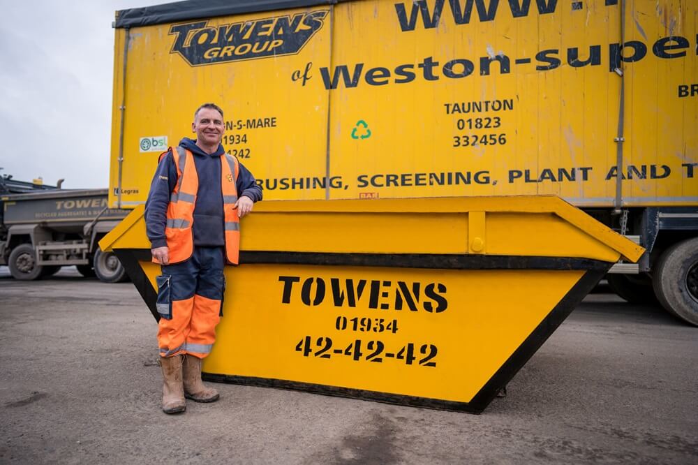 A man in high-visibility orange workwear, including a jacket and pants, stands beside a large yellow skip with the text 'TOWENS' printed on it in black. The skip also displays contact numbers and mentions services such as crushing, screening, plant, and transport. The man appears cheerful and confident, leaning casually against the skip with one hand in his pocket, on what seems to be an industrial or construction site.