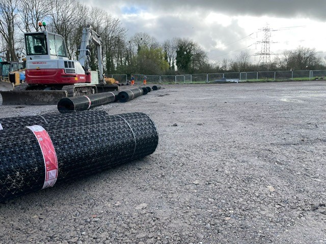 Construction site with rolls of geogrid mesh laid out on compacted ground, with a Takeuchi excavator in the background.