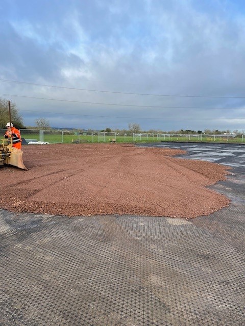 Worker spreading and compacting coarse aggregate over geogrid mesh during sub-base construction on a large site.