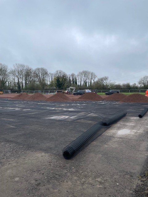 Geogrid mesh being installed on site with compacted layers and piles of aggregate ready for spreading during base construction.