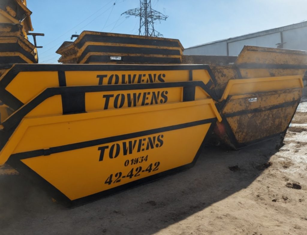 A lineup of various sized yellow Towens skips, each prominently displaying the Towens logo and contact number "01934 42-42-42," arranged in a storage yard. The skips range from smaller to larger capacities, showing varying degrees of wear, under a clear sky with industrial structures in the background.