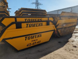 A lineup of various sized yellow Towens skips, each prominently displaying the Towens logo and contact number "01934 42-42-42," arranged in a storage yard. The skips range from smaller to larger capacities, showing varying degrees of wear, under a clear sky with industrial structures in the background.
